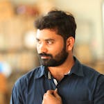 A thoughtful man with a beard and blue shirt posing indoors, displaying a stylish watch.
